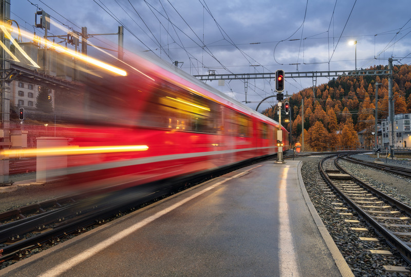 KI generiert: Das Bild zeigt einen schnell vorbeifahrenden roten Zug an einem Bahnhof, umgeben von Herbstlandschaft und Schienen. Im Hintergrund sind Bäume in herbstlichen Farben und der Himmel in der Dämmerung sichtbar.