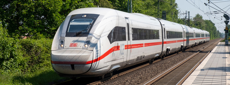 KI generiert: Das Bild zeigt einen modernen Hochgeschwindigkeitszug der Deutschen Bahn, der auf einem Bahngleis fährt. Der Zug ist weiß mit einer roten Linie und fährt bei sonnigem Wetter an einem Bahnhof vorbei.