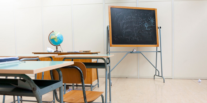 KI generiert: Das Bild zeigt ein leeres Klassenzimmer mit einem Globus auf einem Schreibtisch und einer Tafel, auf der Formeln und Zeichnungen zu sehen sind. Die Tische und Stühle sind auf den Vordergrund gerichtet.