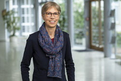 KI generiert: Das Bild zeigt eine Person in einem Innenbereich, die freundlich in die Kamera schaut und einen gemusterten Schal sowie eine Brille trägt. Der Hintergrund ist verschwommen und deutet auf eine moderne, helle Umgebung hin.