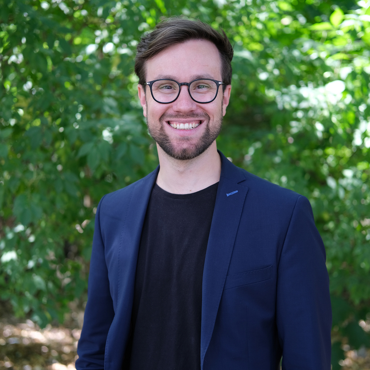 KI generiert: Ein lächelnder Mann mit Brille und Bart steht im Freien vor einer grünen, blättrigen Hintergrundkulisse. Er trägt einen dunkelblauen Blazer über einem schwarzen T-Shirt.