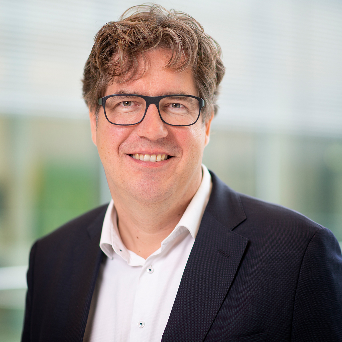 KI generiert: Das Bild zeigt einen lächelnden Mann mit Brille und lockigem Haar in einem Anzug. Der Hintergrund ist verschwommen und lässt auf einen Innenraum schließen.