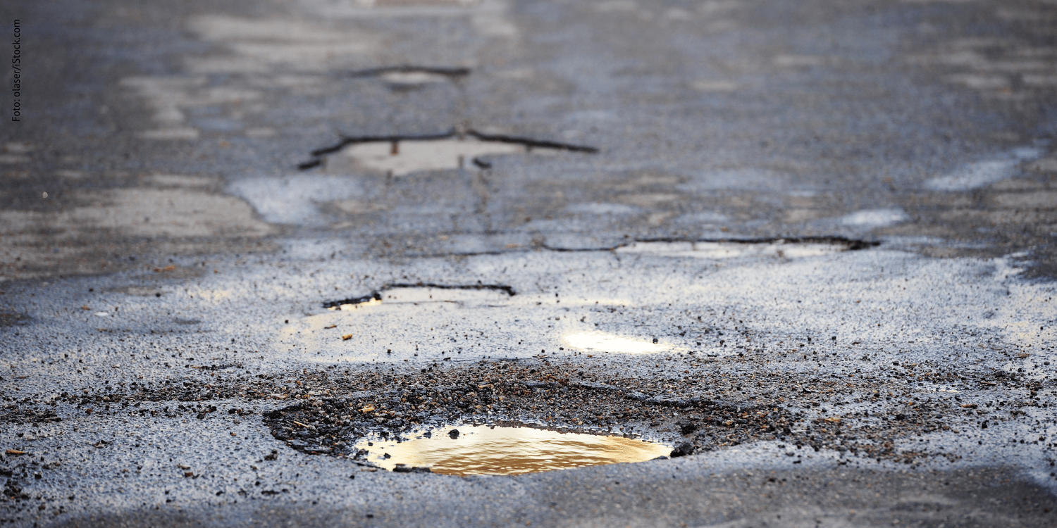 KI generiert: Das Bild zeigt eine beschädigte Straße mit mehreren Schlaglöchern, in denen sich Wasser gesammelt hat. Die Schlaglöcher reflektieren das Licht und verleihen dem Asphalt eine unebene Textur.