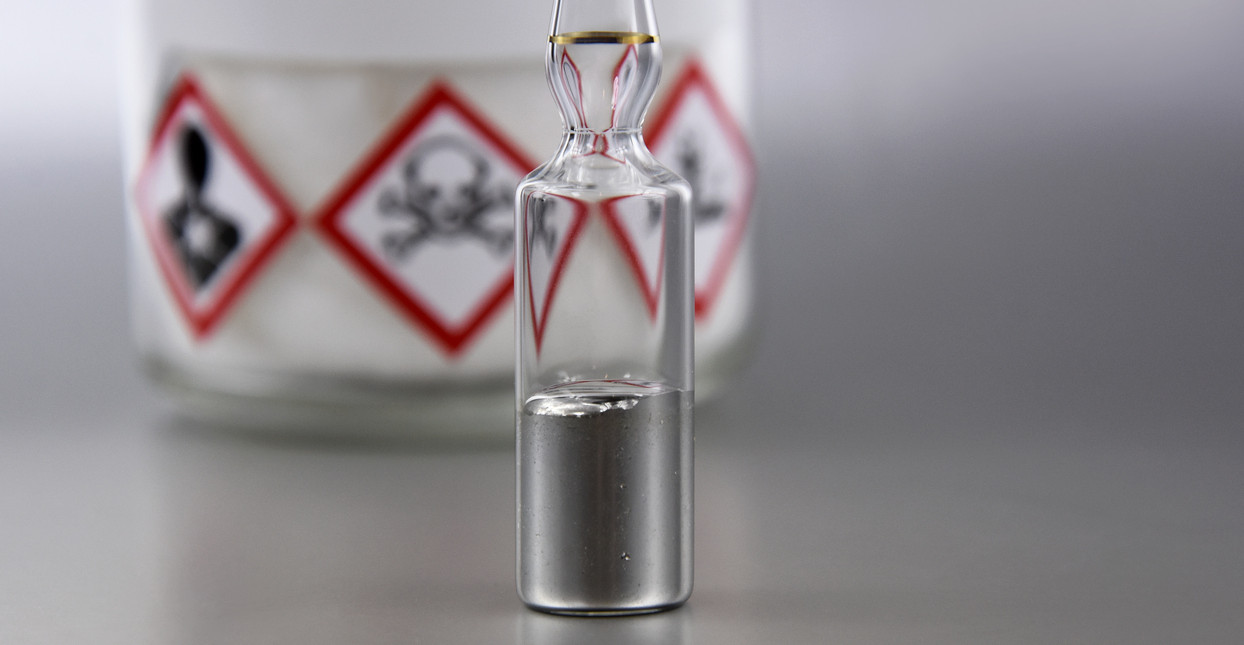 KI generiert: Das Bild zeigt eine Glasampulle, die mit einer silbrigen Flüssigkeit gefüllt ist, vermutlich Quecksilber. Im Hintergrund sind Warnsymbole auf einem Behälter sichtbar, die auf Giftigkeit hinweisen.