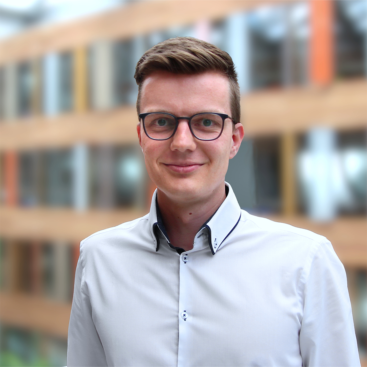 KI generiert: Das Bild zeigt einen Mann, der in einem hellen Hemd lächelt und eine Brille trägt, vor einem unscharfen Hintergrund, der ein modernes Gebäude darstellt. Der Fokus liegt auf dem Porträt des Mannes.