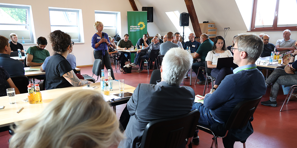 KI generiert: Das Bild zeigt eine Gruppenveranstaltung in einem Raum, bei der mehrere Personen sitzen und einer Frau aufmerksam zuhören, die gerade spricht. Im Hintergrund ist ein Banner mit dem Logo einer politischen Partei zu sehen.