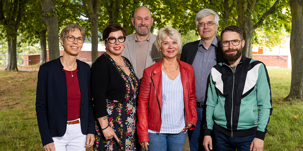 KI generiert: Das Bild zeigt sechs lächelnde Personen, die in einem grünen, von Bäumen umgebenen Park stehen. Sie scheinen sich für ein Gruppenfoto zu positionieren.