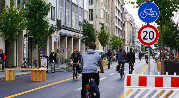KI generiert: Das Bild zeigt eine belebte städtische Straße, die vorwiegend von Radfahrern genutzt wird. Verkehrsschilder weisen auf eine Geschwindigkeitsbegrenzung von 20 km/h und einen Fahrradweg hin.