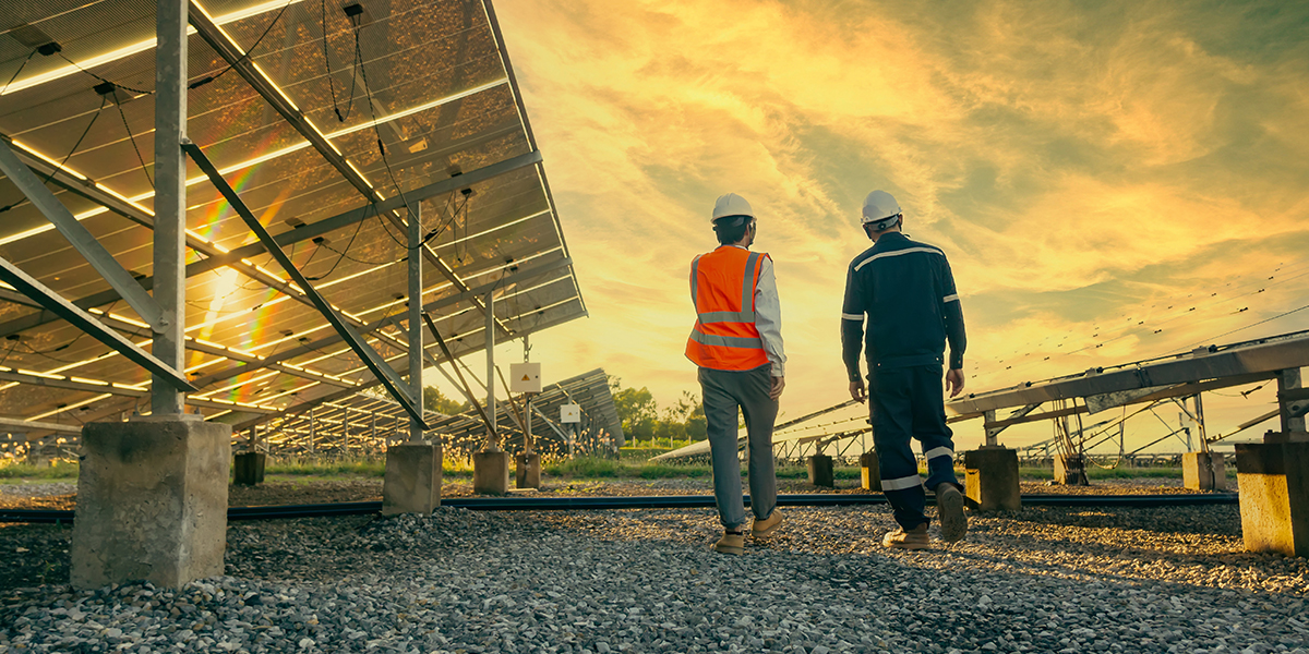 KI generiert: Das Bild zeigt zwei Personen mit Schutzhelmen und Sicherheitswesten, die durch eine Solaranlage gehen. Im Hintergrund sind Sonnenkollektoren und ein malerischer Himmel zu sehen.