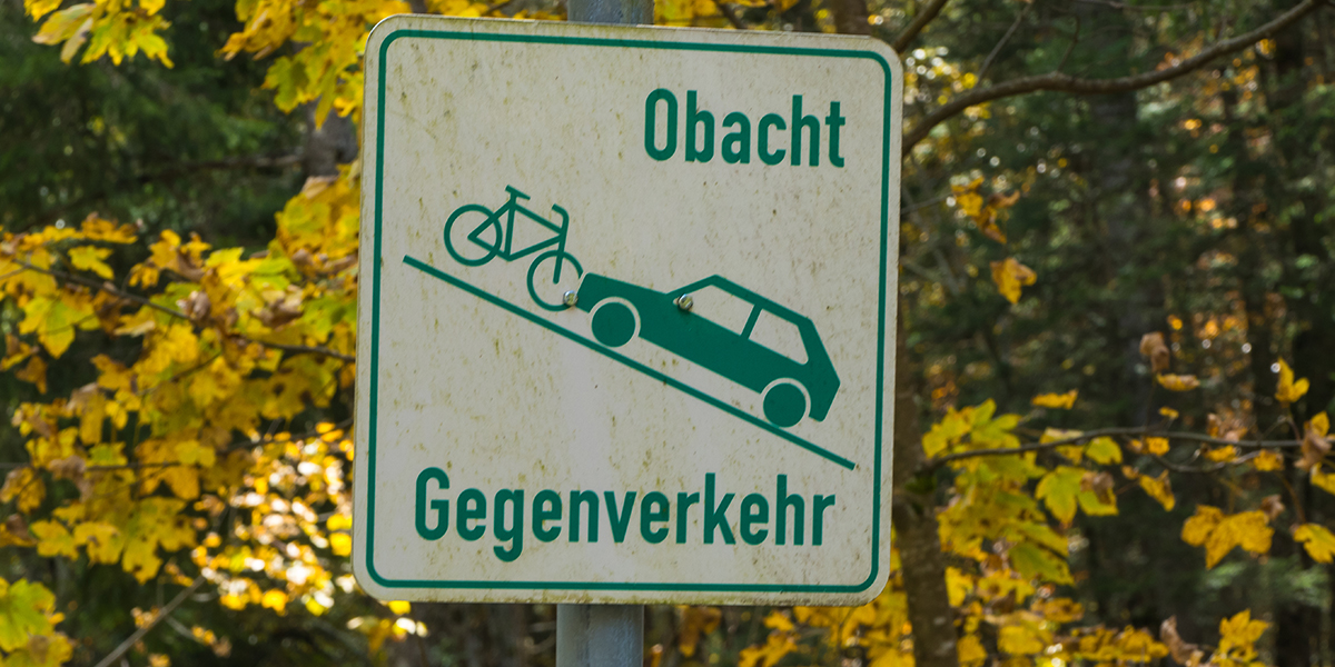 KI generiert: Ein Verkehrsschild warnt vor möglichem Gegenverkehr auf einer schmalen Straße oder einem Weg mit dem Hinweis "Obacht Gegenverkehr" und zeigt ein Auto und ein Fahrrad auf einer Steigung. Die Umgebung ist herbstlich mit gelblichen Blättern.