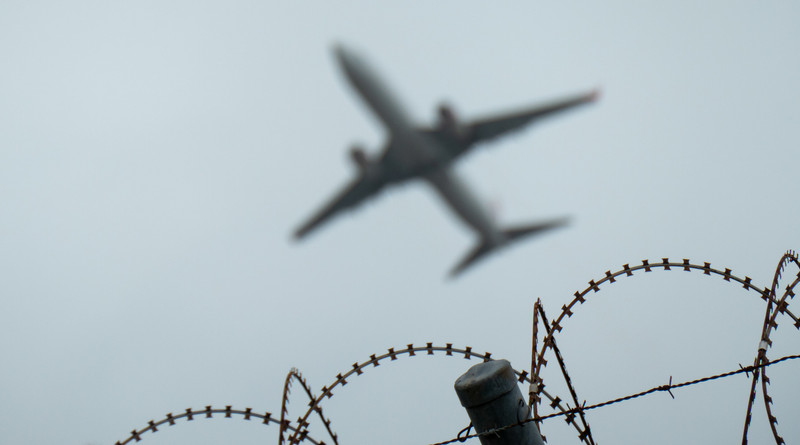 KI generiert: Das Bild zeigt ein unscharf fliegendes Flugzeug über einem Stacheldrahtzaun. Der Fokus liegt auf dem Stacheldraht im Vordergrund, während das Flugzeug im Hintergrund verschwommen ist.