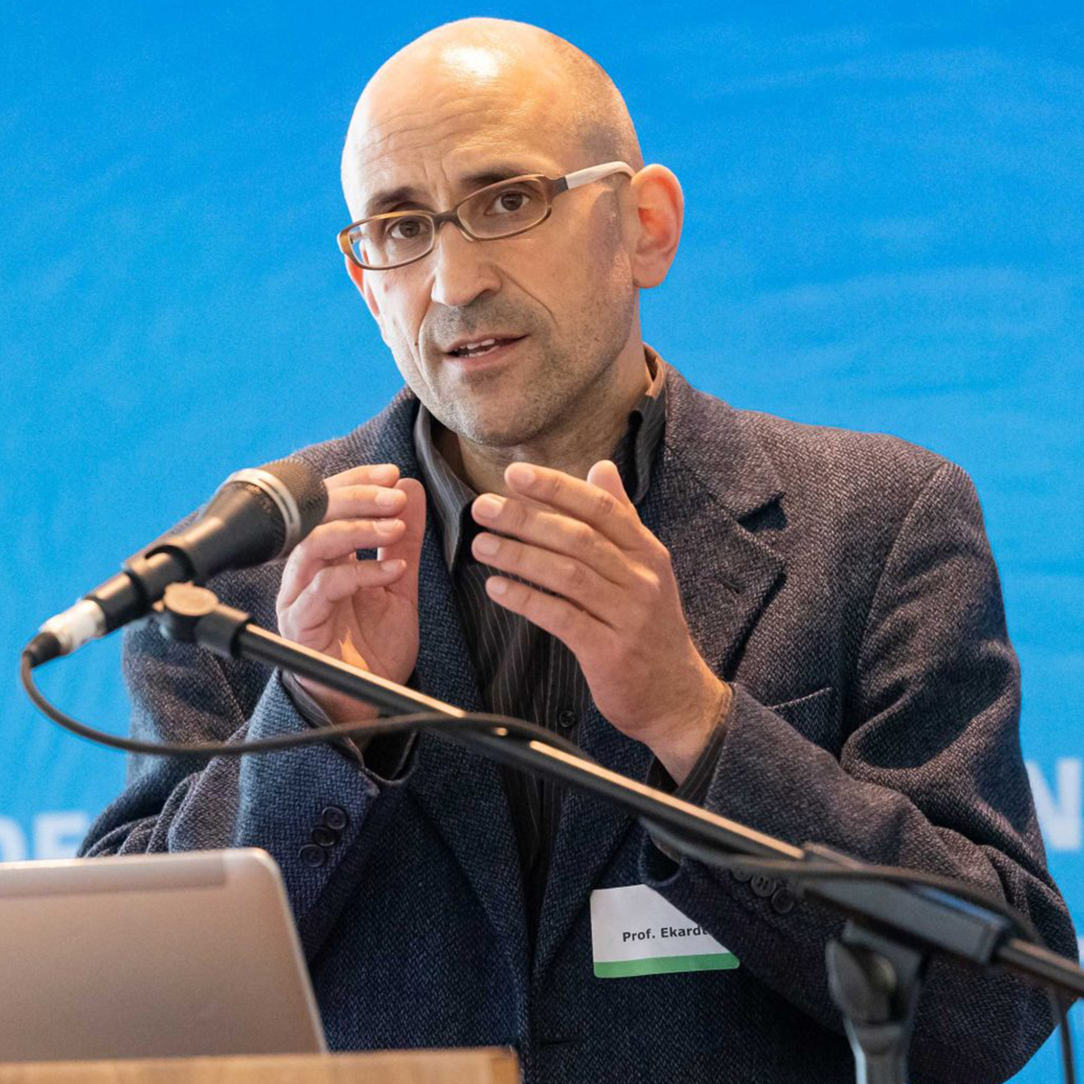 KI generiert: Das Bild zeigt einen Mann, der vor einem Mikrofon steht und spricht, während er seine Hände gestikuliert. Er trägt eine Brille und ist formell gekleidet.
