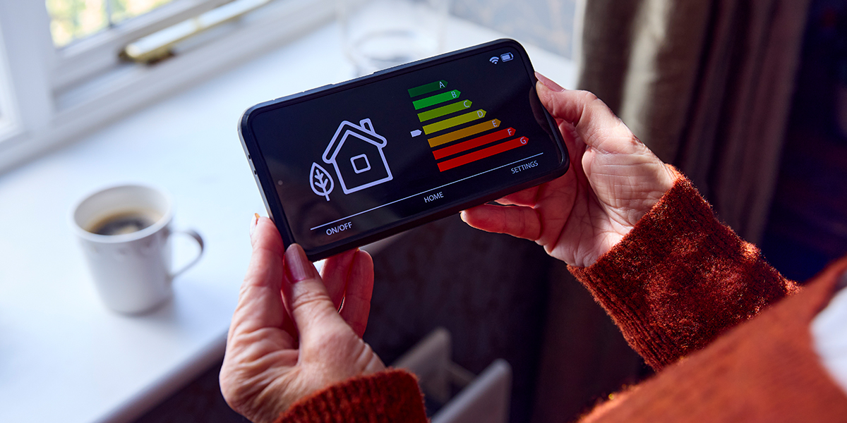 KI generiert: Das Bild zeigt eine Person, die ein Smartphone mit einer App hält, die den Energieverbrauch eines Hauses anhand eines Energieeffizienzdiagramms anzeigt. Im Hintergrund ist ein Fenster und eine Tasse auf einer Fensterbank zu sehen.