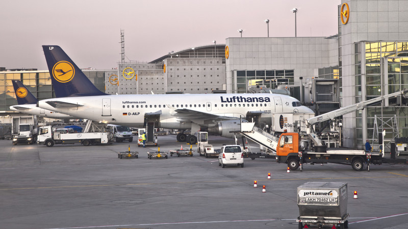 KI generiert: Das Bild zeigt ein Lufthansa-Flugzeug, das an einem Gate eines Flughafens steht. Im Vordergrund sind verschiedene Bodenfahrzeuge und Gepäckwagen zu sehen, die am Flugzeug arbeiten.