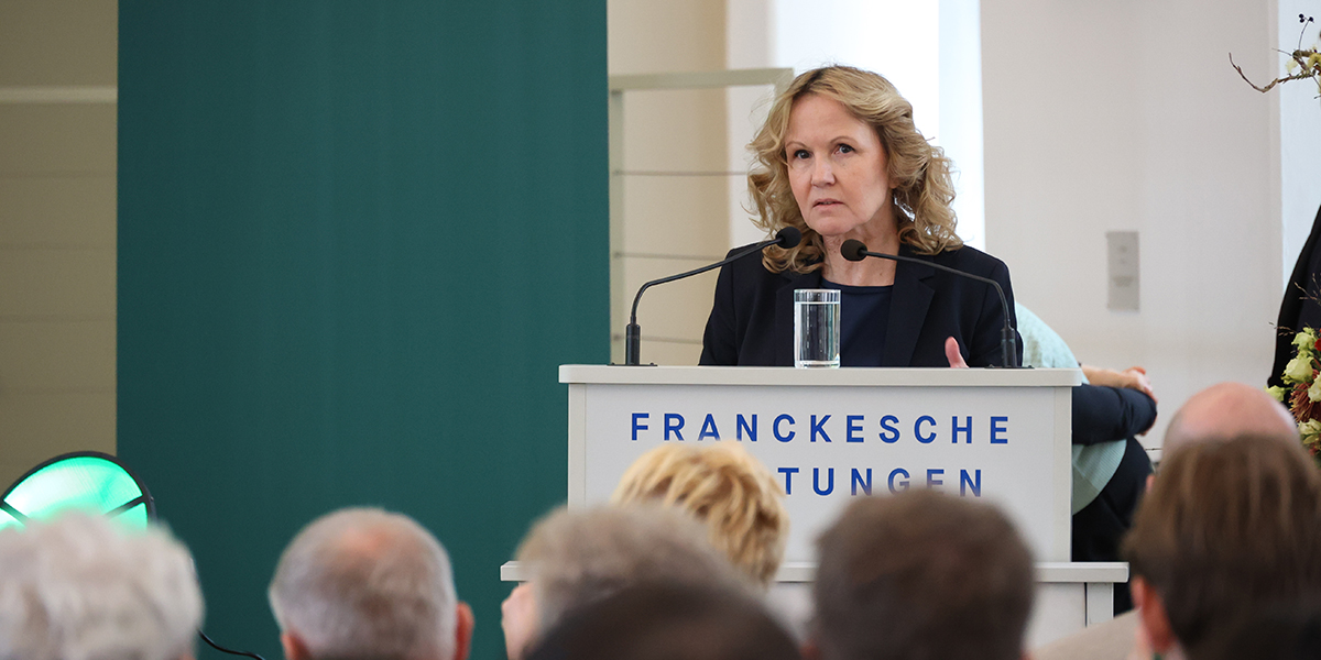 KI generiert: Das Bild zeigt eine Frau, die an einem Rednerpult mit der Aufschrift "Franckesche Stiftungen" steht und eine Ansprache hält. Im Vordergrund sind verschwommen Personen zu erkennen, die ihr zuhören.