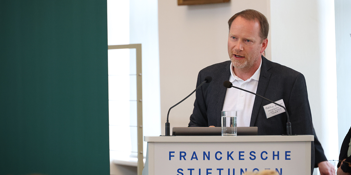 KI generiert: Ein Mann hält eine Rede an einem Podium bei den Franckeschen Stiftungen. Dabei trägt er ein Namensschild und hat ein Glas Wasser vor sich.