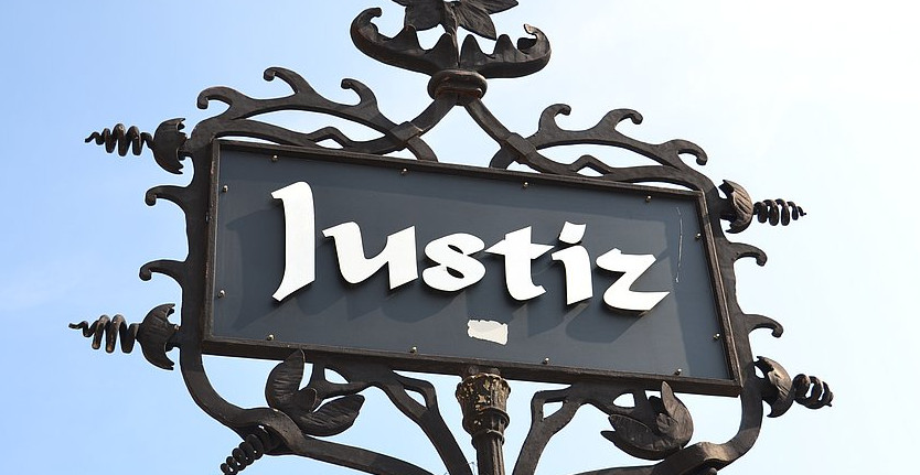 KI generiert: Das Bild zeigt ein schmiedeeisernes Schild mit dem Wort "Justiz" darauf. Der Hauptinhalt ist ein dekoratives Hinweisschild, das möglicherweise auf ein Gerichtsgebäude hinweist.