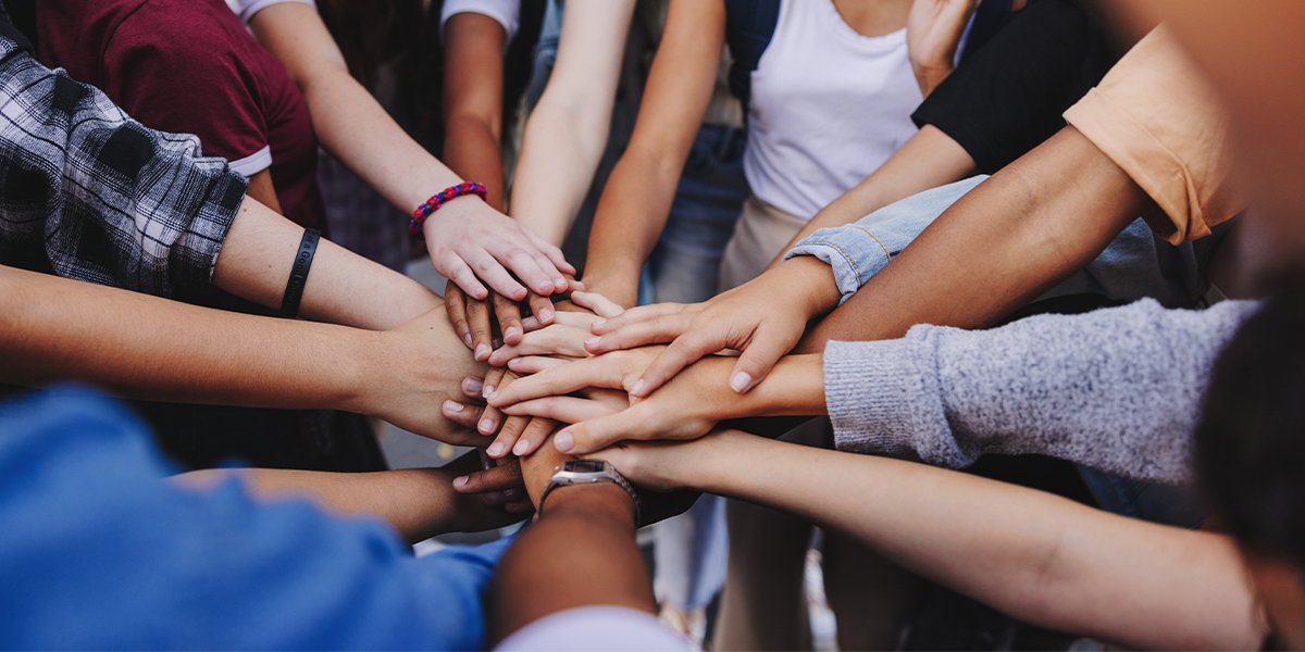 KI generiert: Das Bild zeigt eine Gruppe von Menschen, die ihre Hände in der Mitte übereinanderlegen, was Zusammenhalt und Teamarbeit symbolisiert. Der Fokus liegt auf der Vielfalt der Hände und der gemeinsamen Geste.