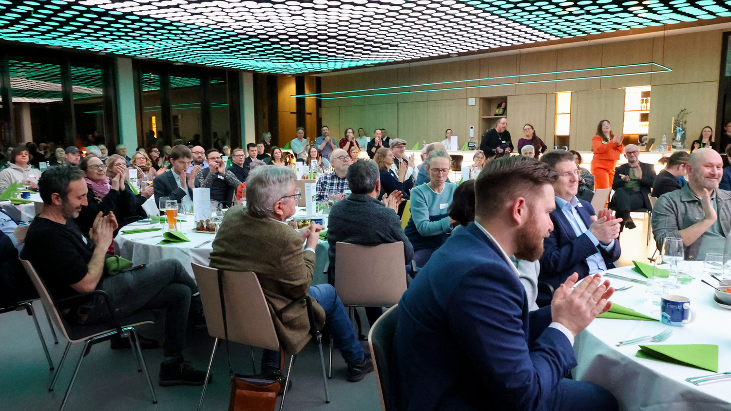 KI generiert: Das Bild zeigt eine größere Gruppe von Menschen, die bei einer Veranstaltung an Tischen sitzen und applaudieren. Der Raum ist modern beleuchtet und die Tische sind dekoriert.