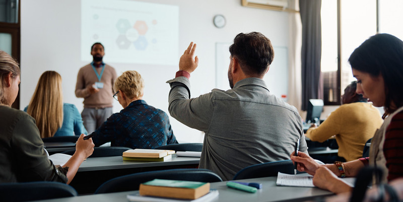 KI generiert: Das Bild zeigt eine Unterrichtssituation, in der ein Dozent vor einer Gruppe von Menschen steht, während eine Person sitzend die Hand hebt, um eine Frage zu stellen. Im Hintergrund ist eine Präsentation zu sehen, die zur Wissensvermittlung eingesetzt wird.