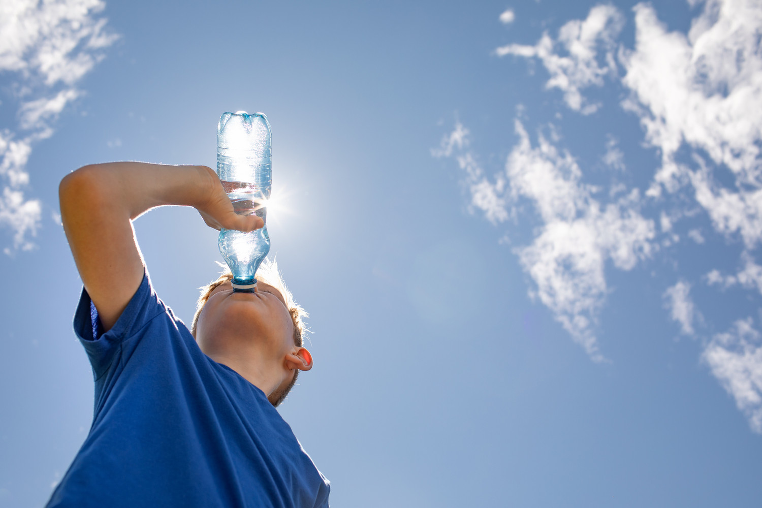 KI generiert: Ein Kind in einem blauen T-Shirt trinkt aus einer Wasserflasche, während es unter einem sonnigen, teilweise wolkigen Himmel steht. Die Sonne scheint hinter der Flasche hervor.