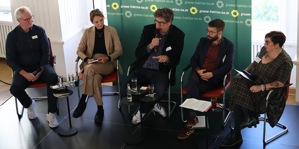 KI generiert: Das Bild zeigt eine Podiumsdiskussion mit fünf Personen, die in Stühlen sitzen und sich unterhalten. Im Hintergrund ist ein Banner mit dem Logo und der Webseite der Grünen-Fraktion Sachsen-Anhalt zu sehen.