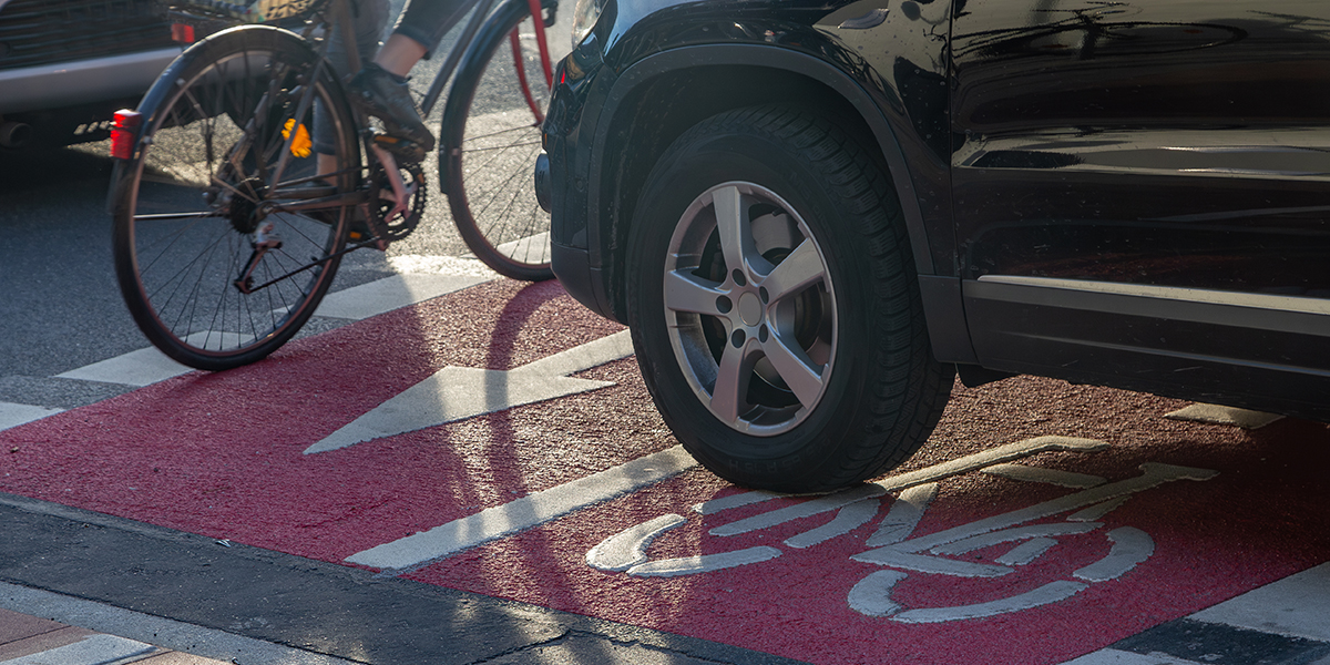 KI generiert: Ein Auto steht auf einem rot markierten Fahrradweg, während ein Fahrradfahrer daran vorbeifährt. Der Fokus liegt auf der Beeinträchtigung des Fahrradbereichs durch das parkende Auto.