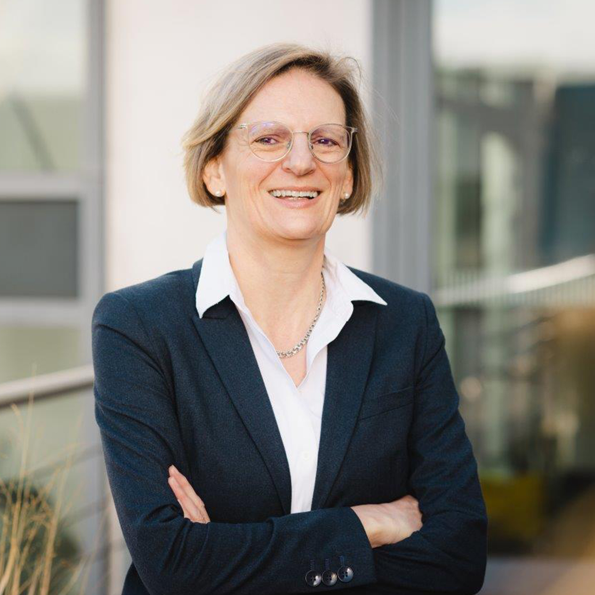 KI generiert: Das Bild zeigt eine lächelnde Person in einem Business-Outfit mit verschränkten Armen vor einem Gebäudeteil im Hintergrund. Der Fokus liegt auf der freundlich und professionell wirkenden Ausstrahlung der Person.