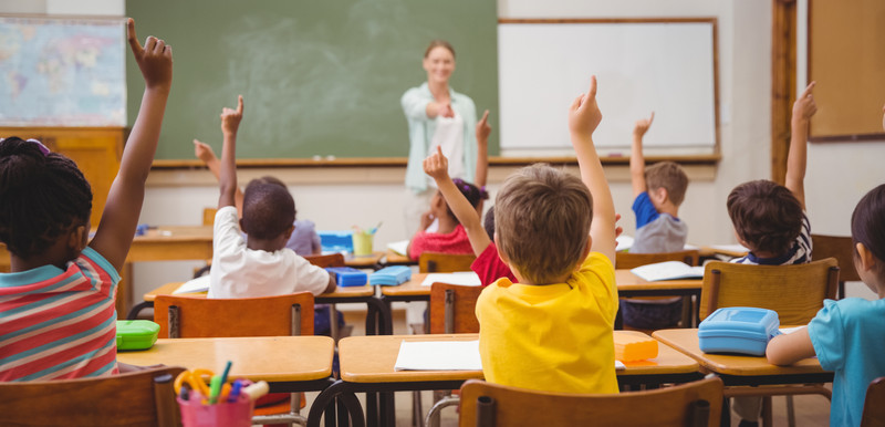 KI generiert: Das Bild zeigt eine Schulklasse mit Kindern, die sich eifrig melden. Im Vordergrund sitzen die Schüler, während im Hintergrund eine Lehrerin an der Tafel steht.