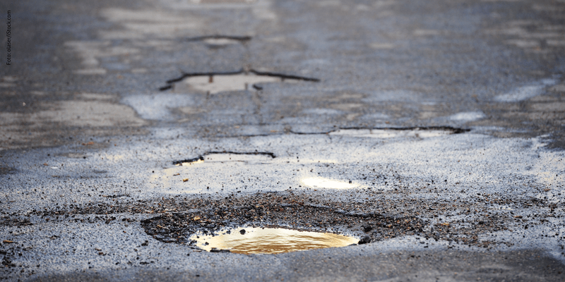 KI generiert: Das Bild zeigt eine Straße mit mehreren Schlaglöchern, in denen sich Wasser gesammelt hat. Die Oberfläche ist sichtlich uneben und beschädigt.