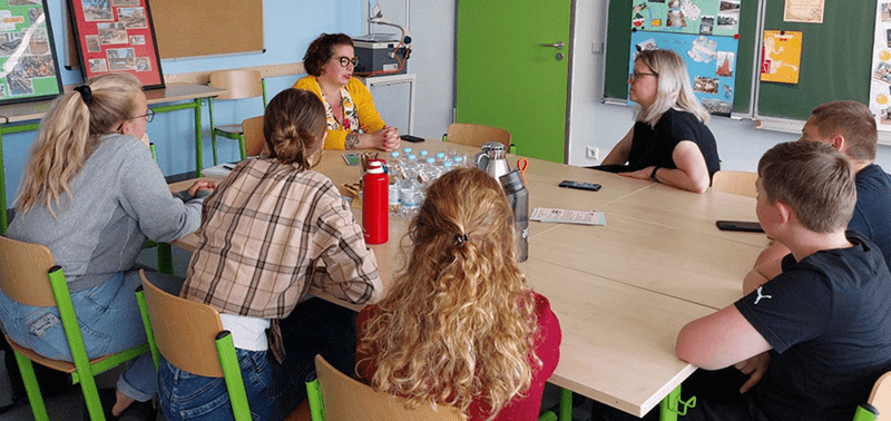 KI generiert: Eine Gruppe von Menschen sitzt in einem Klassenzimmer um einen Tisch und diskutiert. Es scheint sich um ein Gespräch oder eine Besprechung zu handeln.