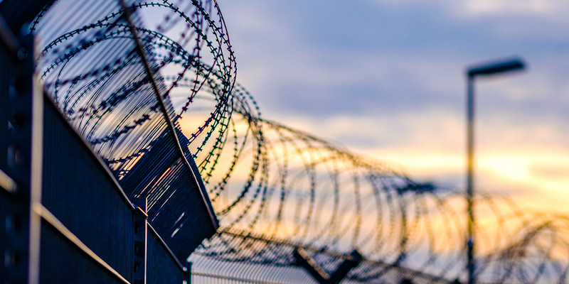 KI generiert: Das Bild zeigt Stacheldraht auf einem Zaun vor einem Sonnenuntergang, wobei der Fokus auf den scharfen Drähten liegt. Der Hintergrund zeigt einen leicht wolkigen Himmel, durch den warmes Licht scheint.