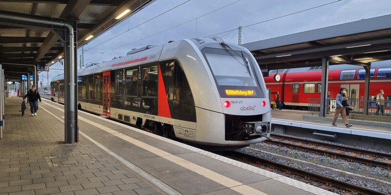 KI generiert: Das Bild zeigt einen modernen Zug am Bahnsteig eines Bahnhofs, mit Passagieren, die entlang des Gleises gehen. Im Hintergrund ist ein weiterer Zug sichtbar.